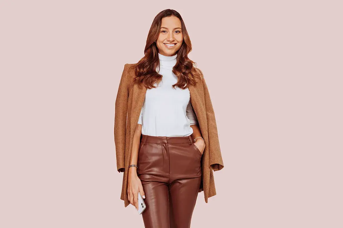 Donna sorridente con un elegante outfit con pantaloni di pelle larghi marroni, dolcevita bianco e cappotto cammello.